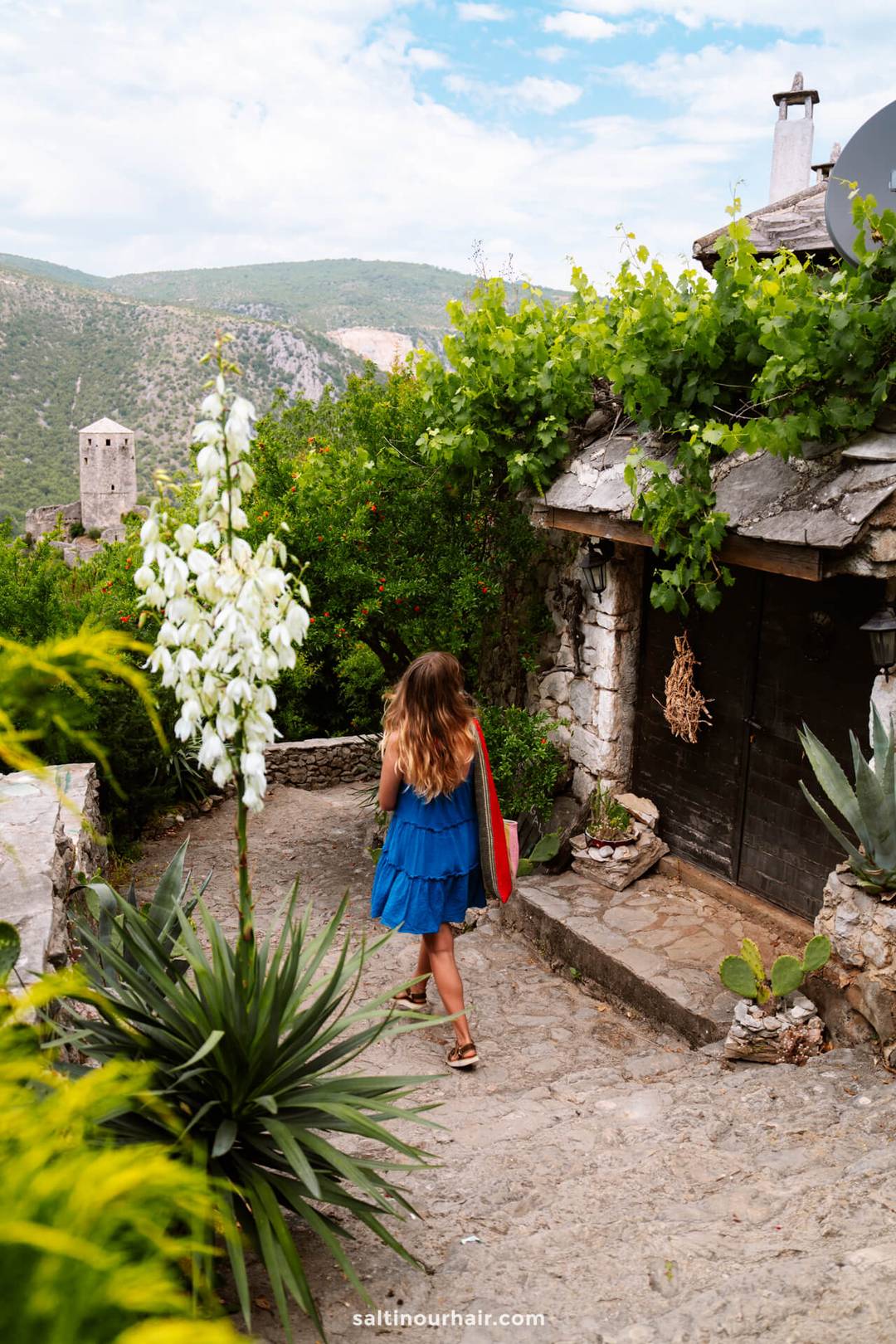 Woman in blue dress walking past a stone house toward Počitelj