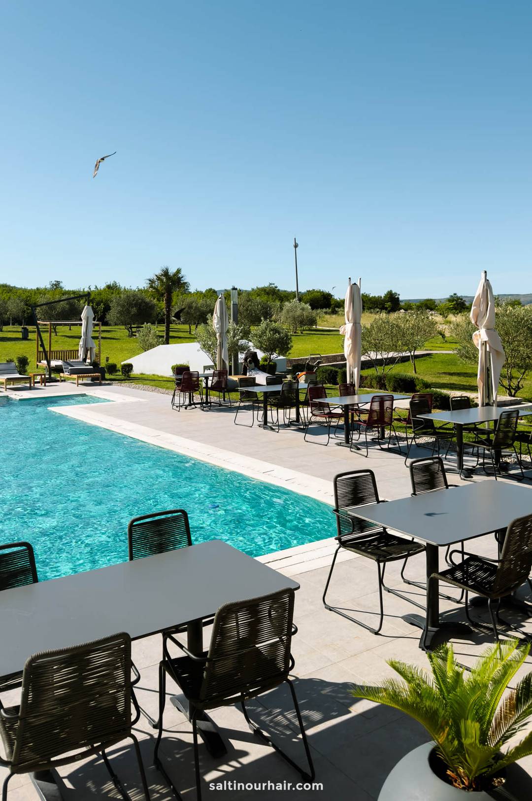 Outdoor pool area with empty tables and chairs on the patio, umbrellas, and a grassy landscape in the background under a clear blue sky&mdash;a relaxing spot among the many things to do in Mostar, Bosnia.