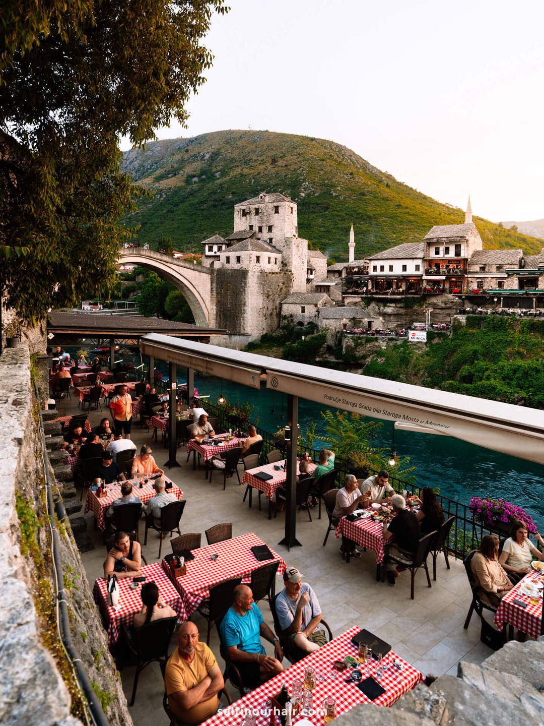 Outdoor restaurant with red-checked tablecloths beside a river, people dining, and a historic stone bridge and old buildings in the background&mdash;a perfect spot to experience one of the top things to do in Mostar, Bosnia.