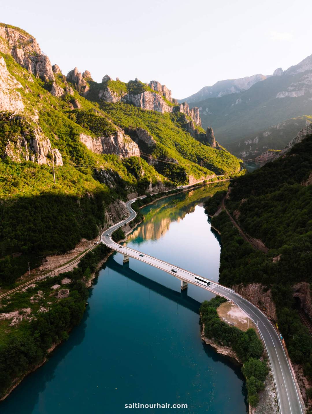 A bridge crosses a winding river surrounded by green mountains and cliffs under clear skies, offering a glimpse of the scenic beauty among the top things to do in Mostar, Bosnia.