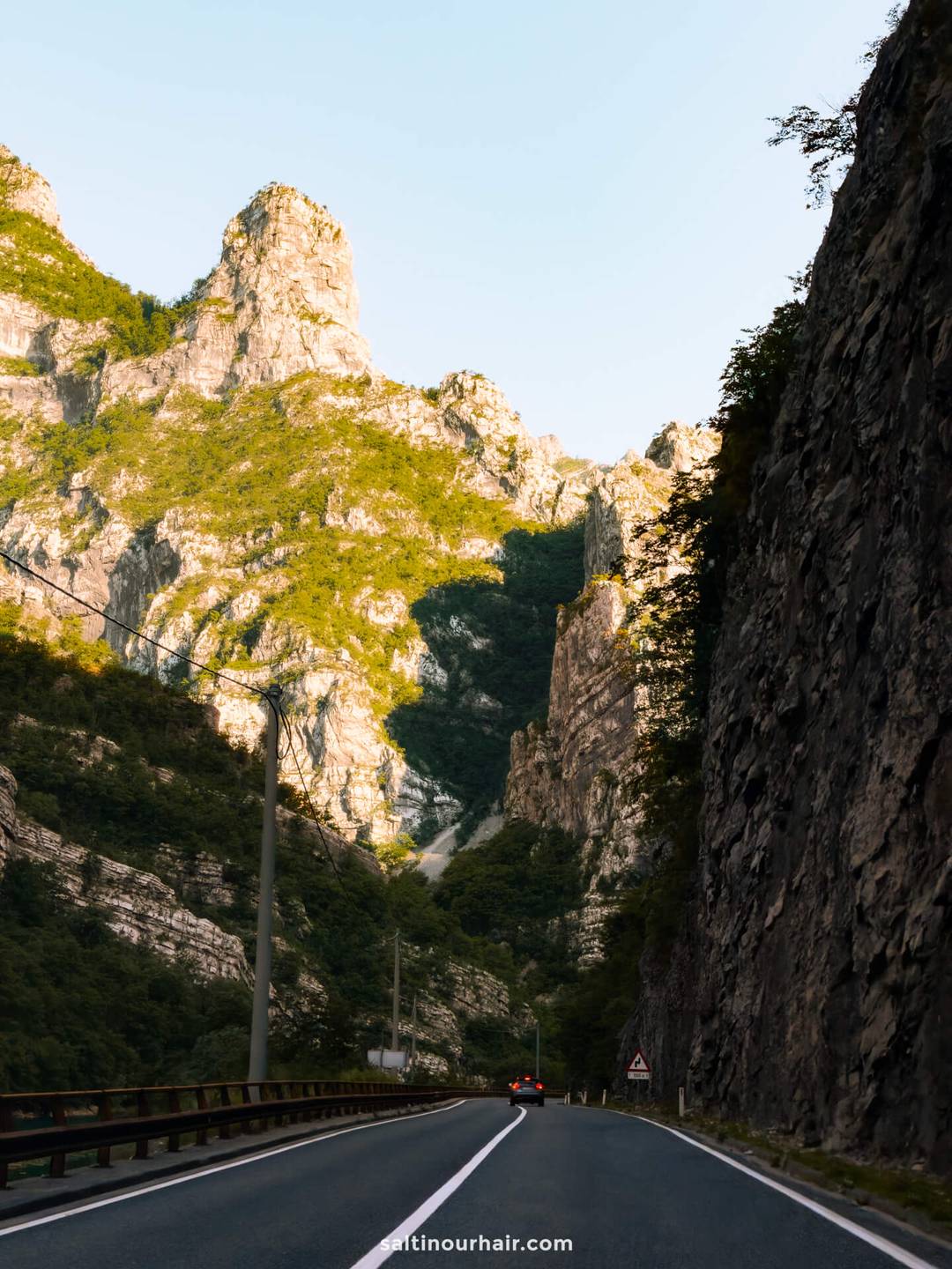 Two-lane road winding through the steep limestone cliffs of Neretva Gorge, with a car ahead and golden sunlight on rocky peaks.