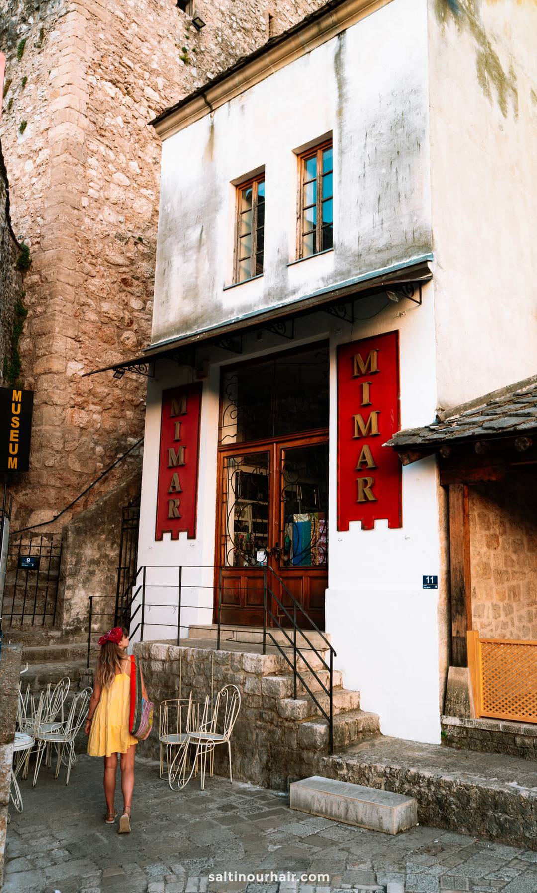 Woman in yellow dress looking up at the Mimar shop entrance in Mostar