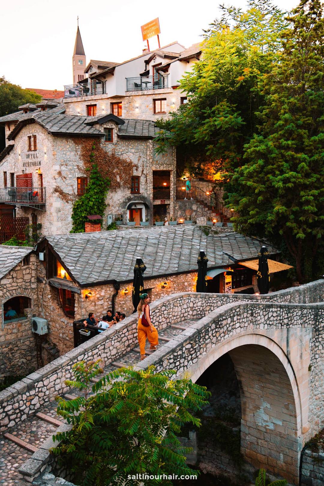 A person walks across a stone arched bridge in Mostar, Bosnia, with historic stone buildings, greenery, and warm outdoor lights in the background&mdash;one of the must-see things to do in Mostar Bosnia.