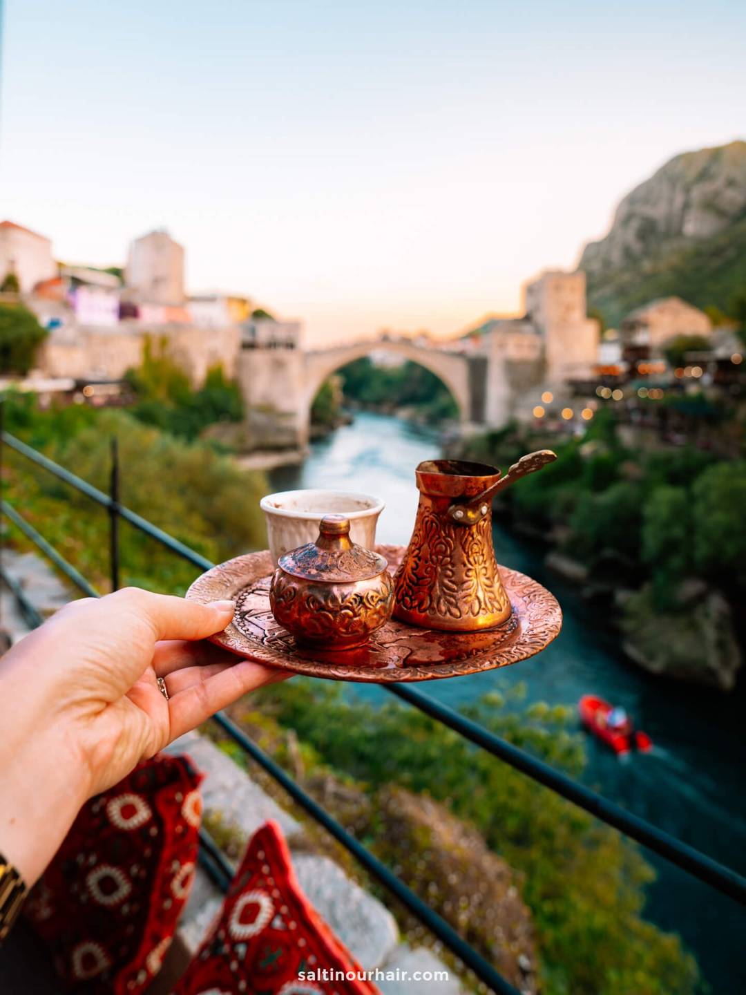A hand holds a tray with a traditional coffee set overlooking a river and an arched stone bridge at sunset&mdash;one of the unforgettable things to do in Mostar, Bosnia.