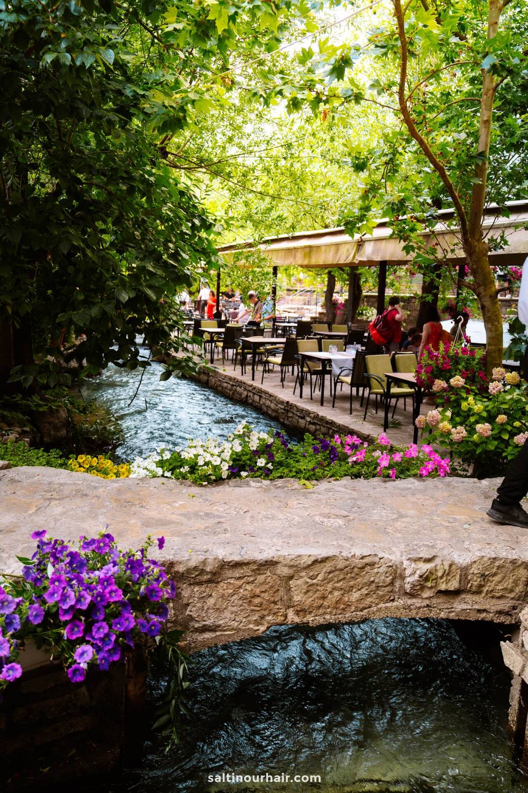 Outdoor riverside caf&eacute; terrace beside the Buna River at Blagaj, Mostar, Bosnia, lined with colorful flowers and shaded trees.