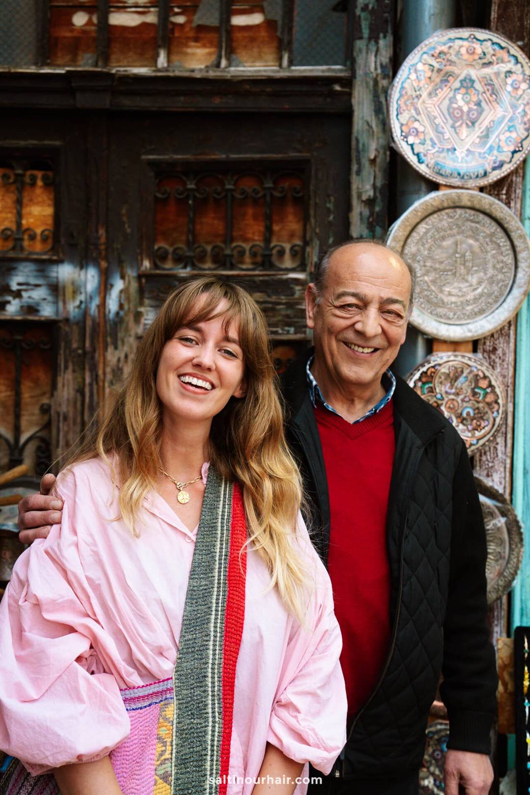 A woman and a man stand side by side, smiling, in front of a rustic wooden door decorated with ornate metal plates&mdash;a perfect moment while exploring Sarajevo things to do.