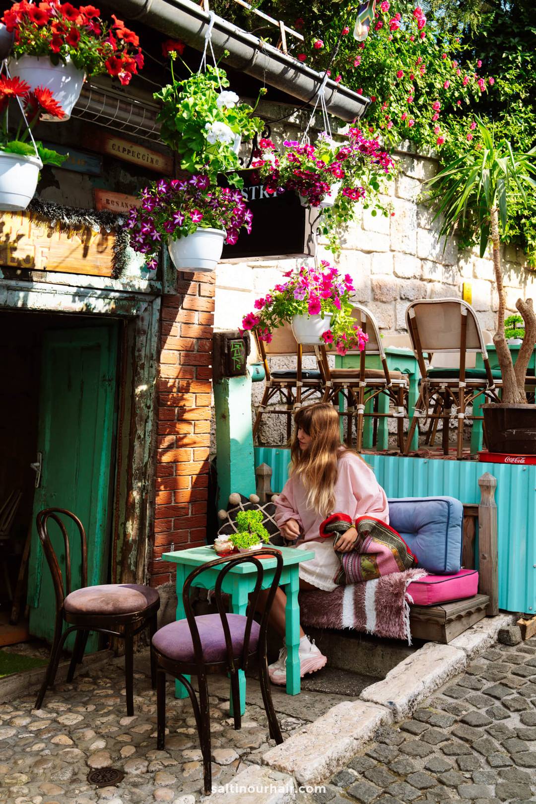 A woman sits at a turquoise table in a colorful outdoor caf&eacute; with vibrant flowers, hanging plants, and rustic d&eacute;cor on a cobblestone street&mdash;capturing one of the charming Sarajevo things to do.