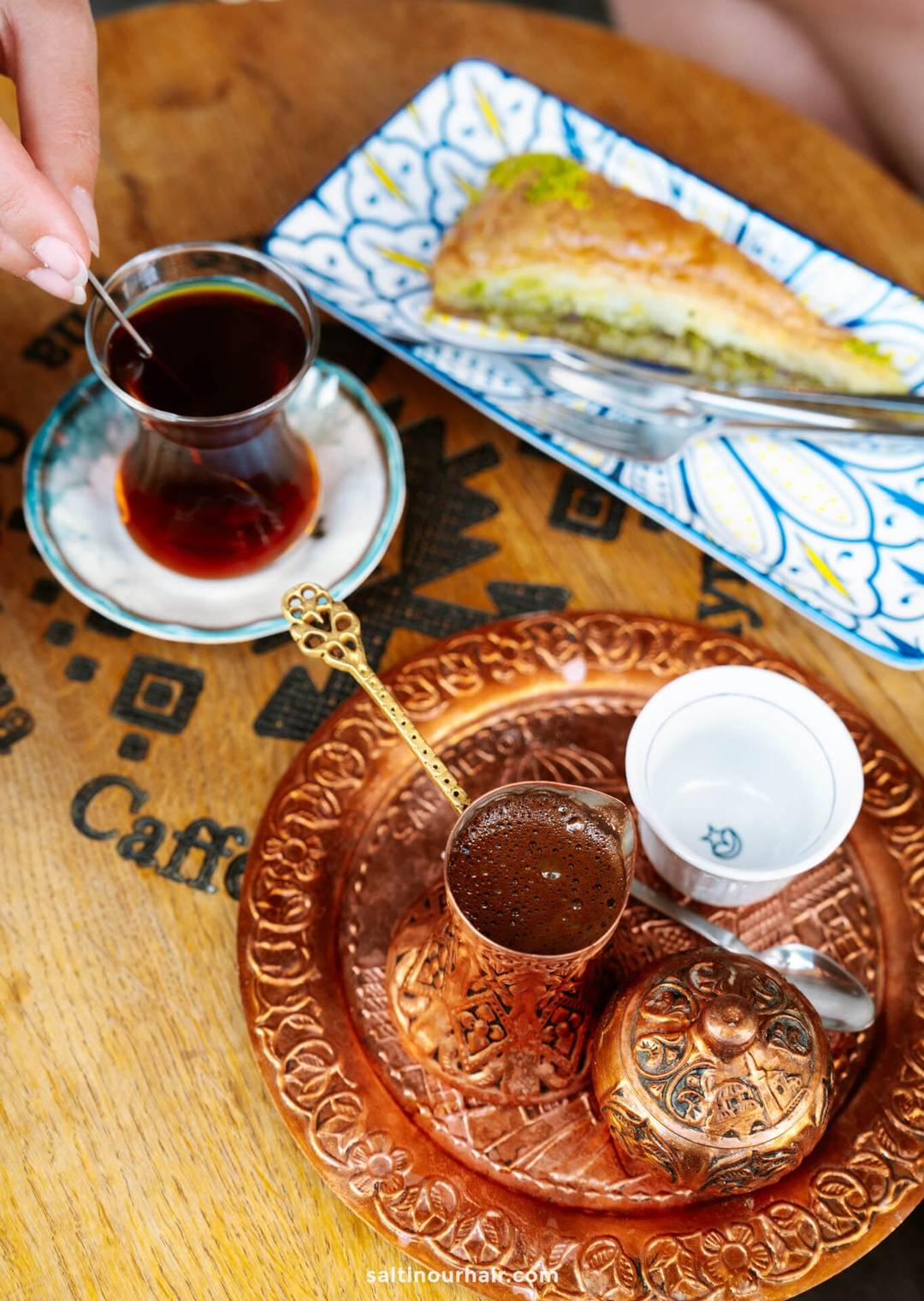 A hand holds a glass of tea next to a plate of baklava and a copper tray with Turkish coffee&mdash;a classic experience among Sarajevo things to do&mdash;on a wooden table.