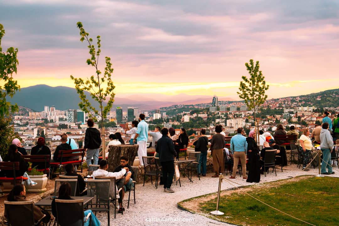A group of people gather at an outdoor caf&eacute; overlooking a cityscape at sunset, with mountains in the distance under a cloudy sky&mdash;one of the best Sarajevo things to do for both locals and visitors.