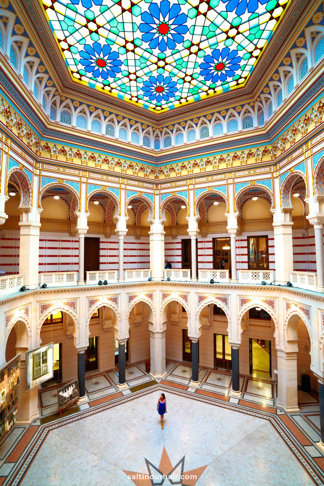 A person stands in the center of an ornate, multi-story hall with arched balconies and a colorful stained-glass ceiling&mdash;an architectural gem among Sarajevo things to do.