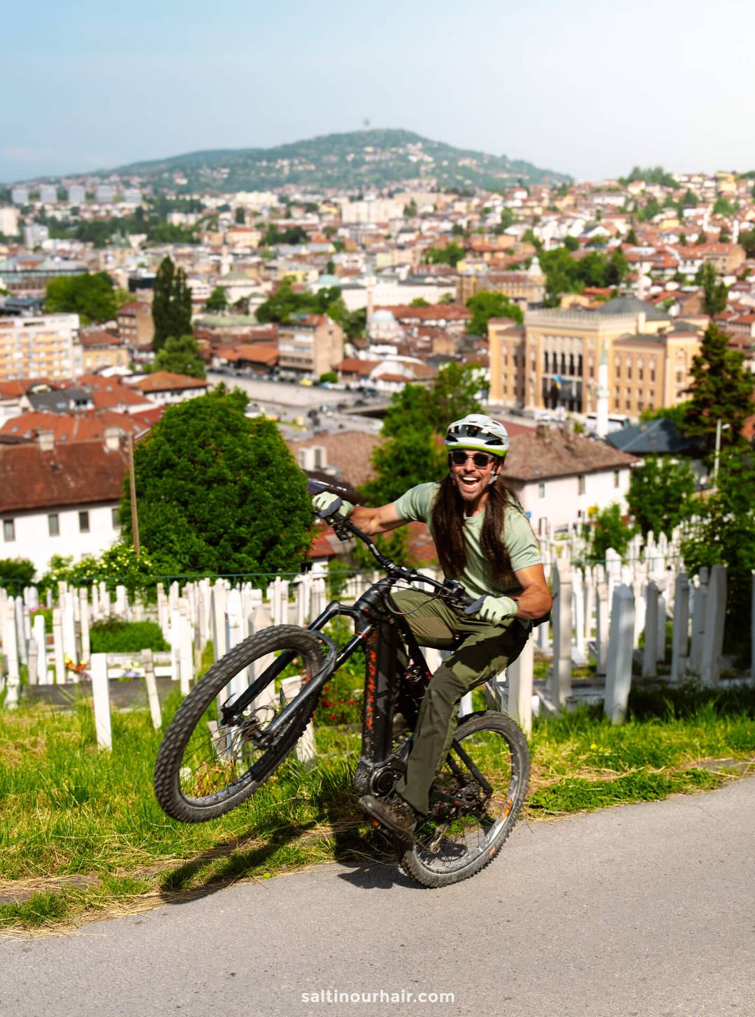 A person wearing a helmet performs a wheelie on a bicycle near a hillside cemetery, with a cityscape and hills in the background&mdash;capturing one of the unique Sarajevo things to do for adventurous visitors.