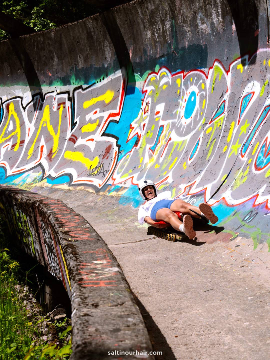 A person wearing a helmet rides a street luge down a graffiti-covered concrete track on a sunny day, showcasing one of the exciting Sarajevo things to do.