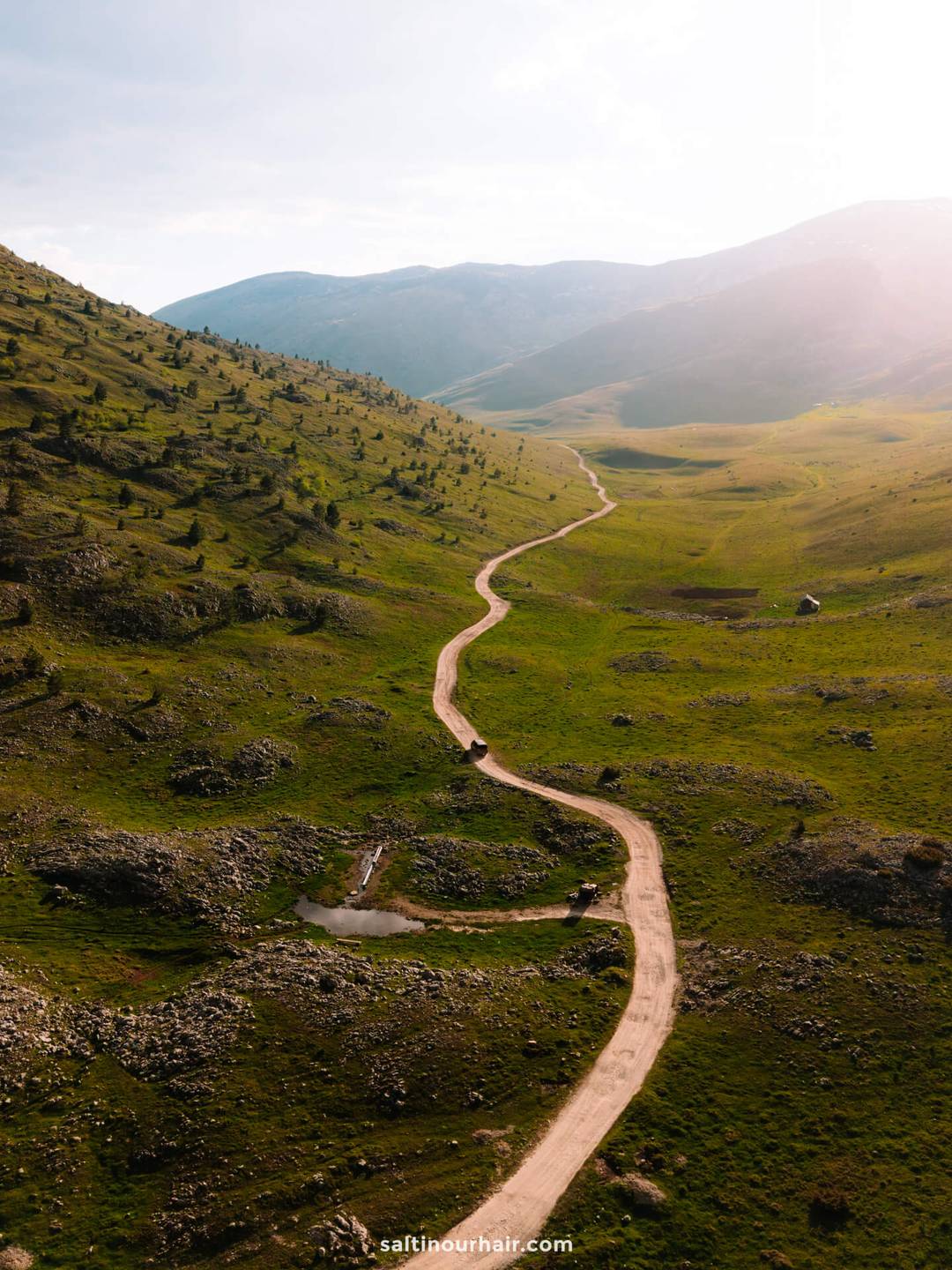 A winding dirt road cuts through a green, hilly landscape under a bright sky with scattered sunlight&mdash;one of many scenic views you