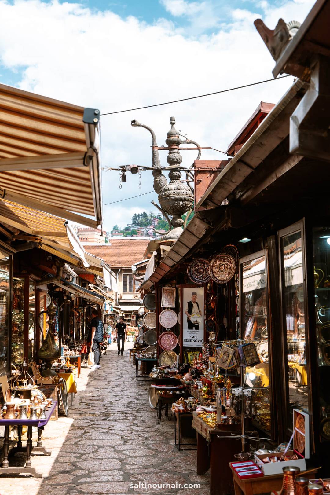 A narrow outdoor market street in Sarajevo, lined with shops selling souvenirs, ceramics, and metalware&mdash;perfect for exploring if you