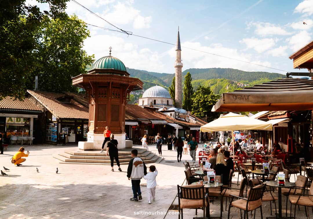 Outdoor plaza with people walking and sitting at caf&eacute; tables, wooden fountain in center&mdash;one of the top Sarajevo things to do&mdash;mosque with dome and minaret in background, surrounded by trees and mountains under a blue sky.