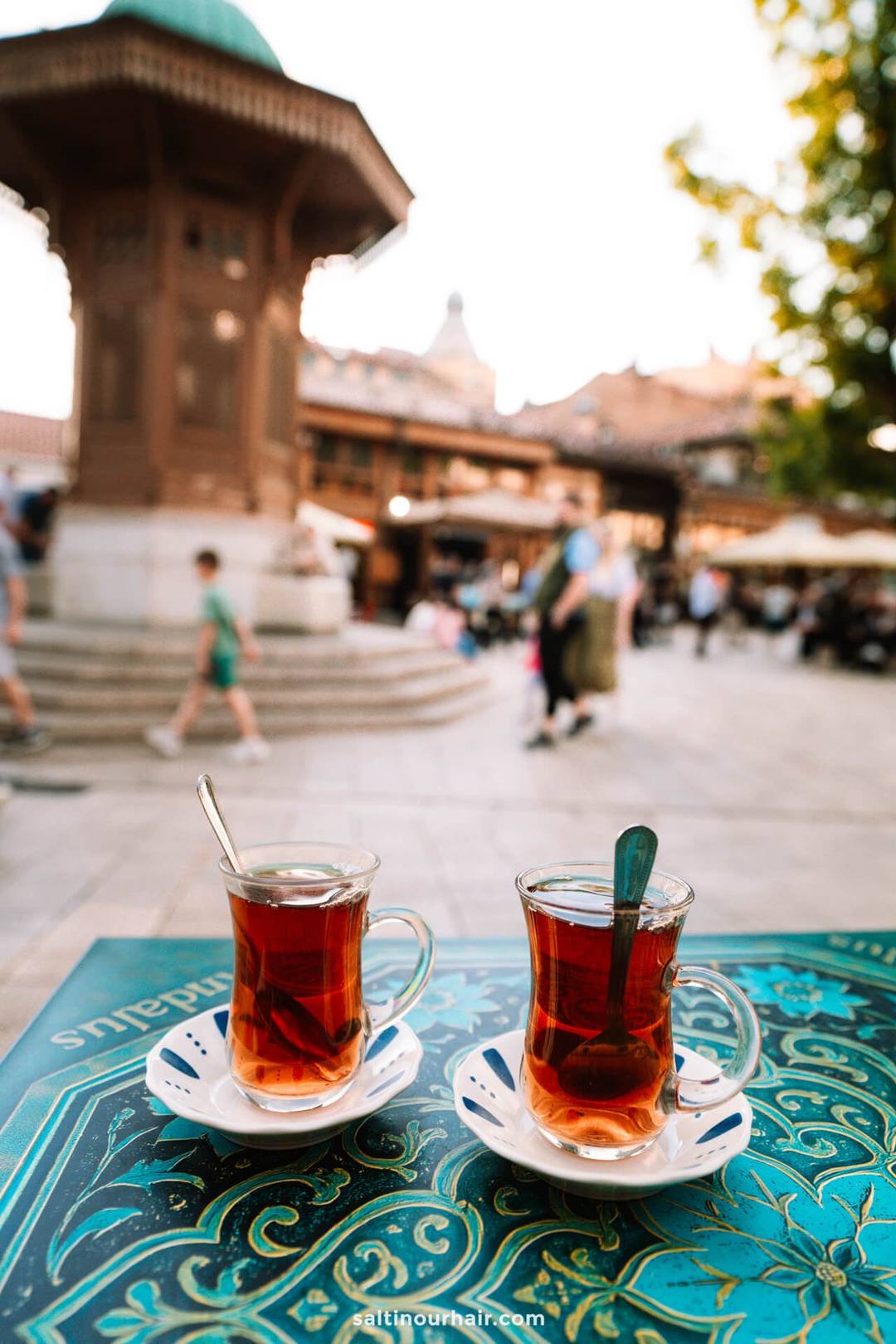 Two glasses of tea with spoons sit on a patterned table in an outdoor square, while people stroll by and a historic fountain rises in the background&mdash;a perfect snapshot of Sarajevo things to do.