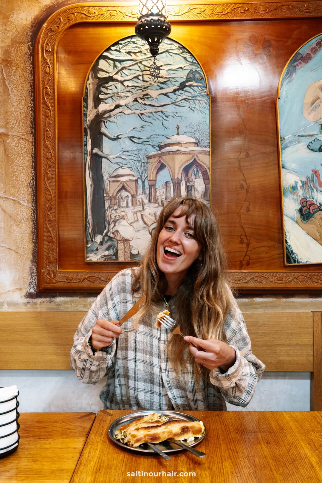 A woman sits at a wooden table in a restaurant, smiling while holding a fork and knife over a plate of pastry&mdash;a delicious highlight among Sarajevo things to do. Framed artwork decorates the wall behind her.
