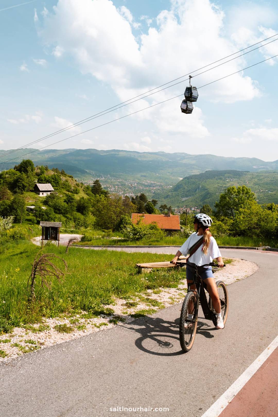 A person wearing a helmet rides a bicycle on a winding path through a green landscape, with cable cars visible overhead and hills in the background&mdash;one of many Sarajevo things to do for outdoor enthusiasts.