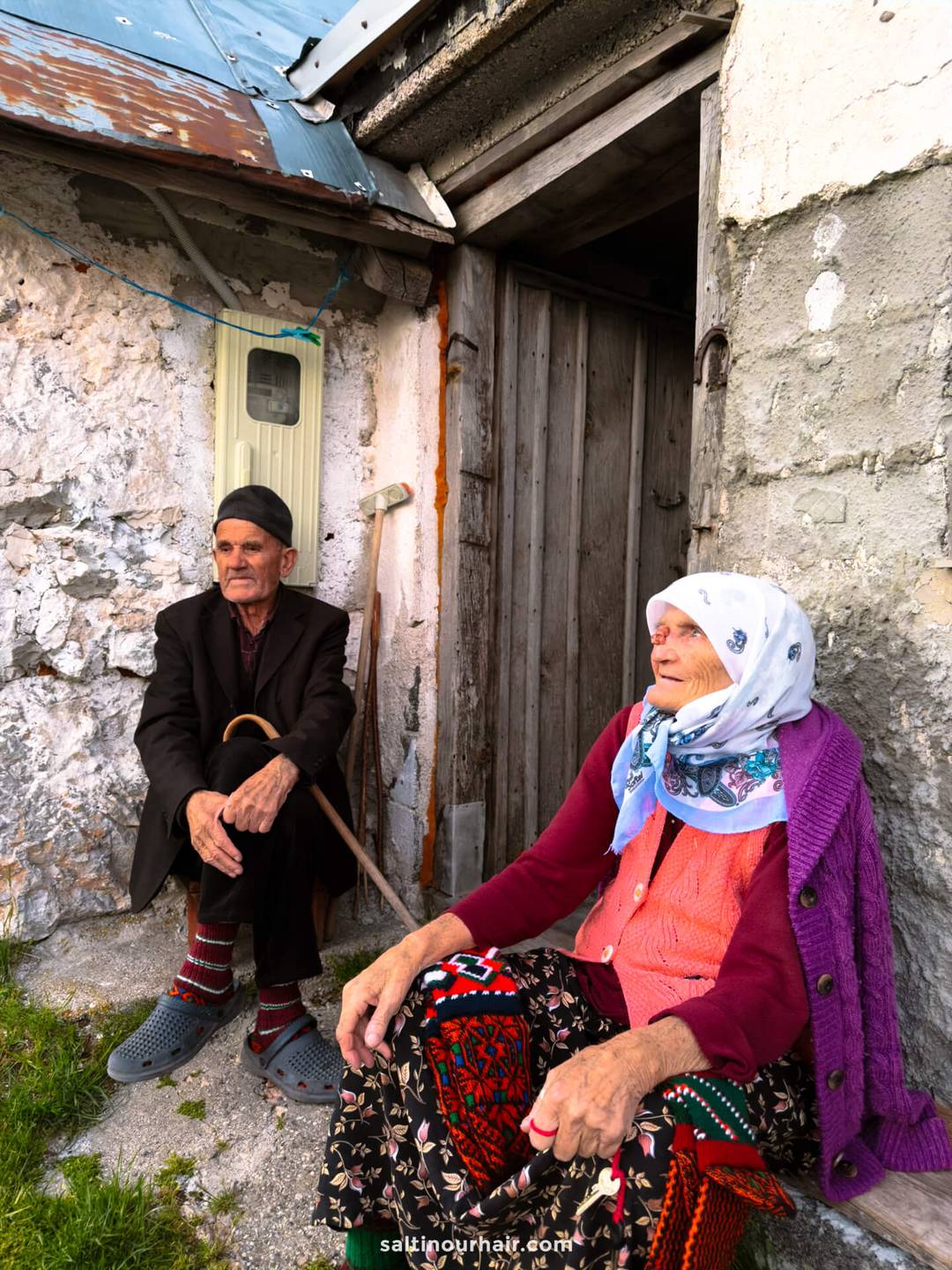An elderly man and woman sit outside a weathered stone building in Sarajevo; the man holds a cane, and both wear traditional clothing with head coverings&mdash;offering an authentic glimpse into Sarajevo things to do and local culture.