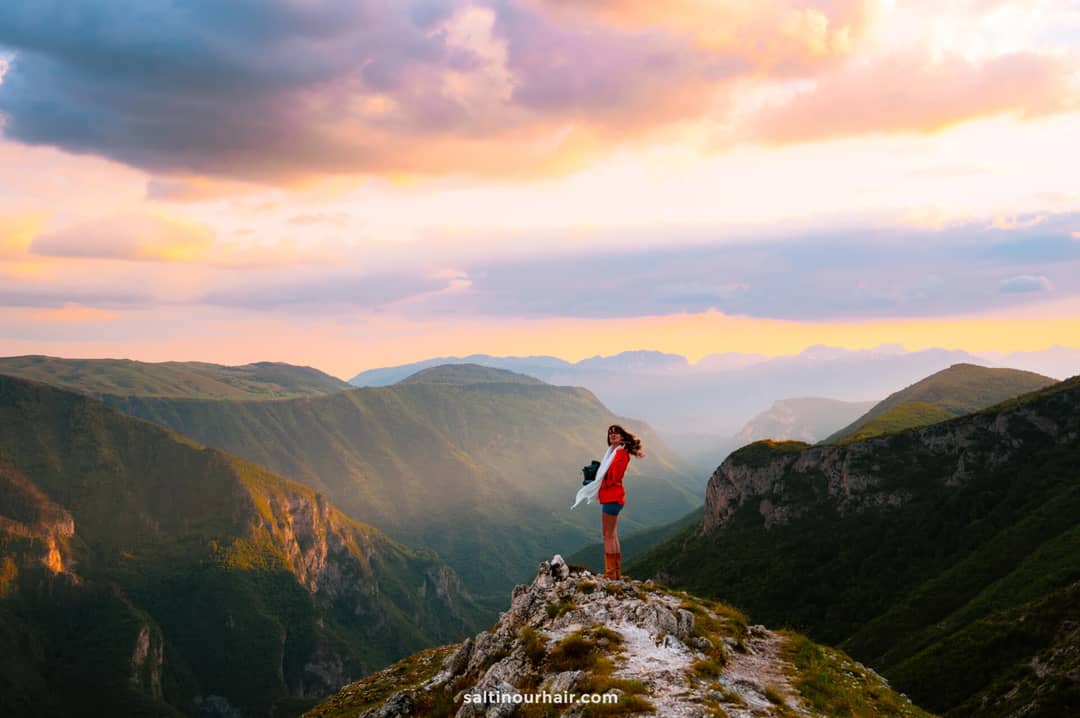 A person stands on a rocky peak overlooking a vast mountain landscape at sunset, with colorful clouds in the sky&mdash;an inspiring scene reminiscent of Sarajevo things to do for adventure and nature lovers.