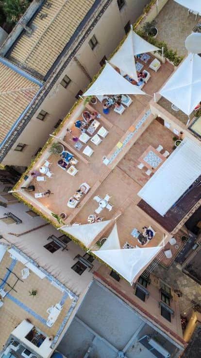 Aerial view of BADIA NUOVA’s rooftop terrace in Trapani, with people sitting at tables, surrounded by buildings and white shade sails overhead.