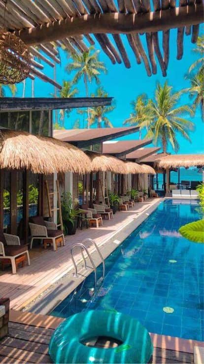 Outdoor pool with blue water at Banig Beach Resort, surrounded by lounge chairs, thatched-roof cabanas, tropical plants, and palm trees—enjoy the clear blue sky in the heart of El Nido.