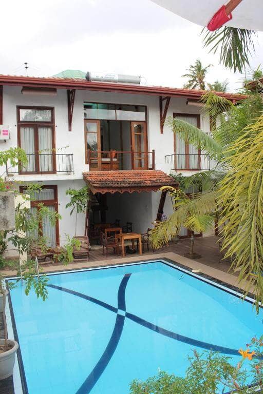 Villa Kapuru in Negombo is a two-story white house with wooden accents, featuring balconies and large windows overlooking a rectangular swimming pool surrounded by lush plants.