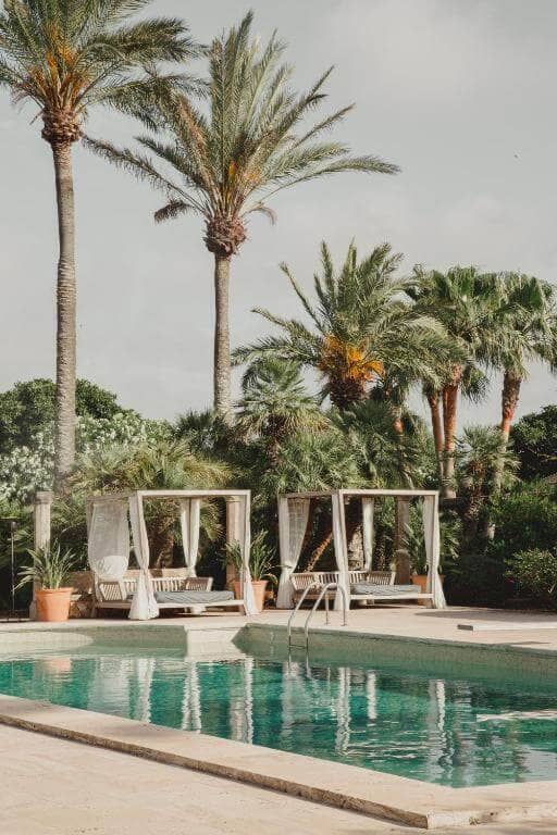 A swimming pool with clear water is in front of two canopy daybeds, surrounded by tall palm trees and lush greenery at Cal Reiet, a holistic retreat in Santanyi.