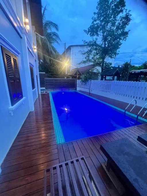 A rectangular queenshouse-lp swimming pool with blue lights is surrounded by a wooden deck, lounge chairs, a white fence, and sits adjacent to the house at dusk.