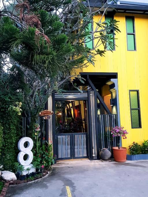 A yellow two-story building with green windows, a metal gate entrance, potted plants, and a large white number 8 sign by the entrance—Baan Baimai Ayutthaya is a welcoming Ayutthaya guesthouse perfect for travelers.