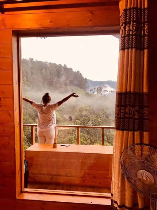 A person stands on a wooden balcony with arms outstretched, overlooking the misty forested landscape of Ella—a true green nature paradise—as seen from inside a room with wood-paneled walls and patterned curtains.