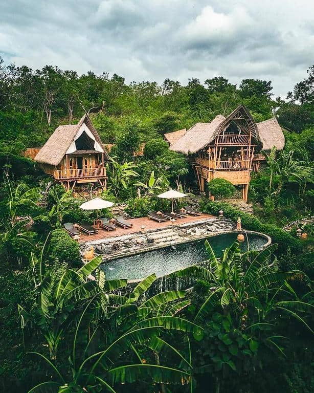 Three thatched-roof wooden villas at Green Suites Penida are surrounded by lush greenery, overlooking a stone-edged pool with lounge chairs and white umbrellas under a cloudy sky.