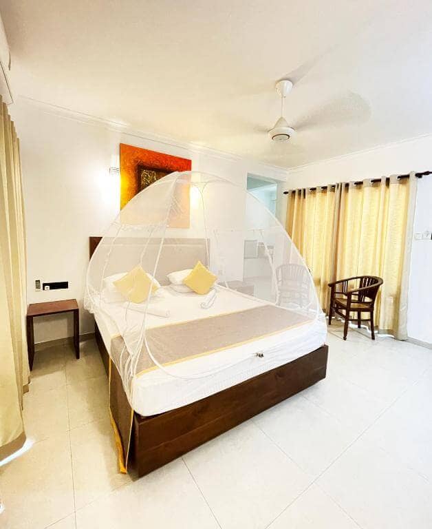 A bright hotel room in Dambulla with a double bed covered by a white mosquito net tent, two chairs, a ceiling fan, and large windows with beige curtains overlooks a stunning rock arch formation.
