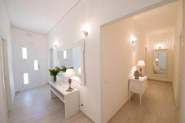 A bright, modern hallway with light-colored walls and floors, decorated with mirrors, lamps, and vases of flowers on white tables, inspired by the serene elegance of Donna Maria Favignana.