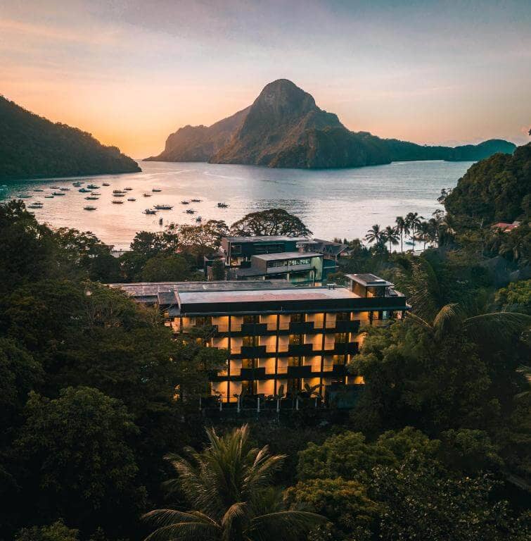 A modern multi-story hotel sits among lush trees in El Nido, with a bay, anchored boats, and a mountain peak in the background at sunset—just steps from the vibrant spirit of the Funny Lion.