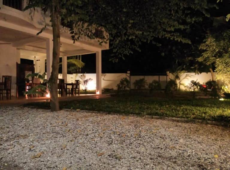 A nighttime view of a patio with tables and chairs, surrounded by a garden and trees in Mirissa, softly illuminated by outdoor lights and accompanied by soothing nature sounds.