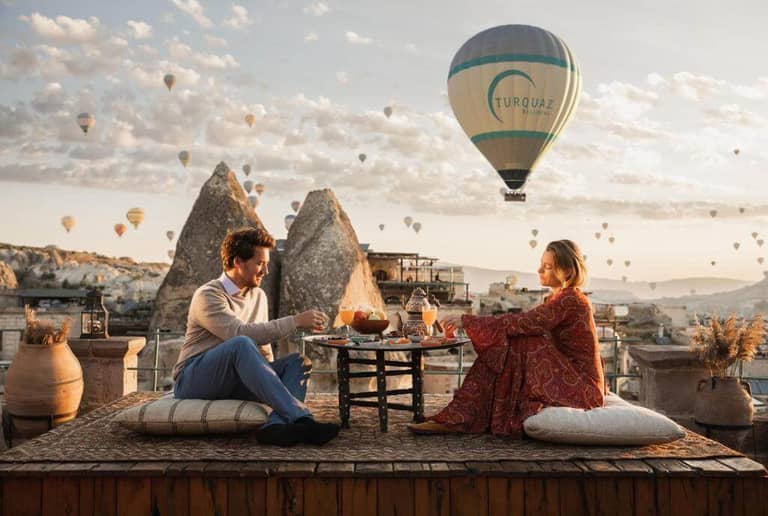 A man and woman sit on cushions at a low outdoor table at Sultan Cave Suites, sharing a meal as hot air balloons rise over the rocky landscape at sunset.