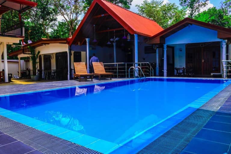 Outdoor swimming pool surrounded by lounge chairs, tiled deck, and several small buildings with red and brown roofs at Sevonrich Dambulla, set amidst lush trees.