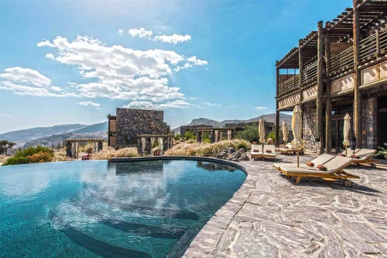 Infinity pool with lounge chairs and umbrellas on a stone patio, adjacent to a rustic building, with mountains and blue sky in the background.