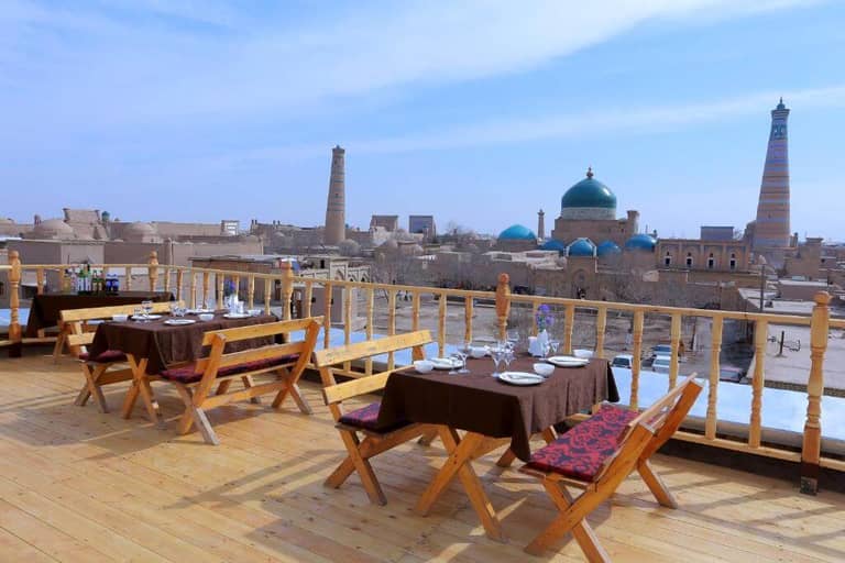 Outdoor restaurant terrace with wooden tables and chairs overlooking Khiva’s ancient cityscape, where domes and minarets rise under a clear sky—an atmospheric setting reminiscent of the legendary Arkanchi.