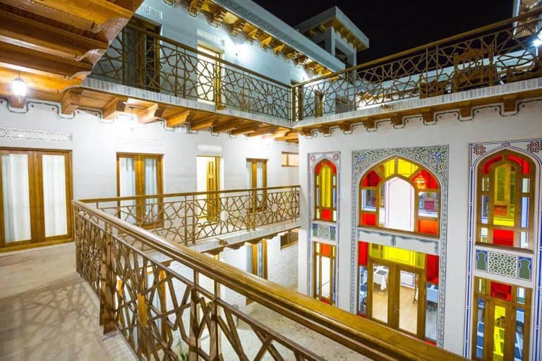 Night view of a traditional Bukhara courtyard building with wooden railings, brightly lit windows, and stained glass detailing on the upper floor—an inviting scene in the heart of Volida.