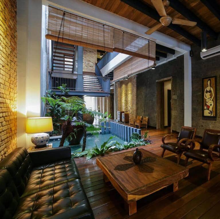 Modern living room with wooden floors, indoor plants, a small pond, leather furniture, exposed brick walls, and an open staircase—evoking the chic vibe of an art gallery in Malacca.