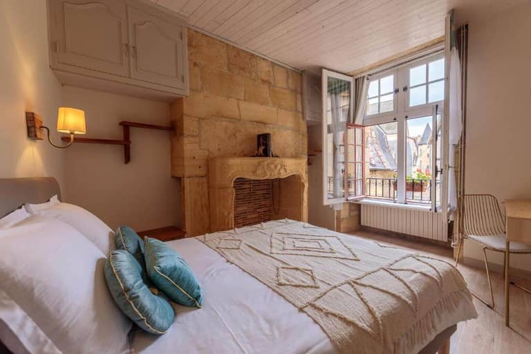 A cozy bedroom at le-lys-sarlat with a double bed, two blue cushions, a decorative fireplace, wooden shelves, a desk and chair, and an open window offering rooftop views.