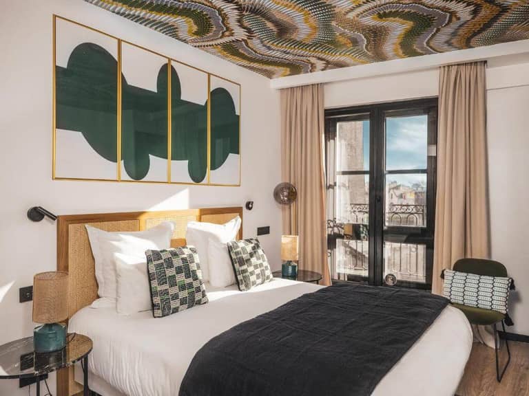 Modern hotel room in La Rochelle featuring a double bed, patterned pillows, wall art, glass side tables, and floor-to-ceiling windows with beige curtains and a decorative patterned ceiling—perfect for your Monde getaway.