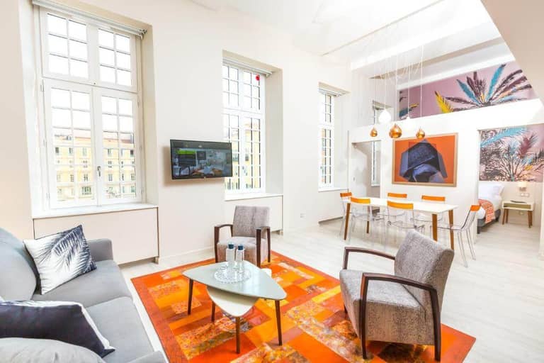 Bright living and dining area with large windows, modern furniture, orange accents, wall art, and a mounted TV—perfect for a stylish stay at Palais Saleya, a boutique hotel in Nice.