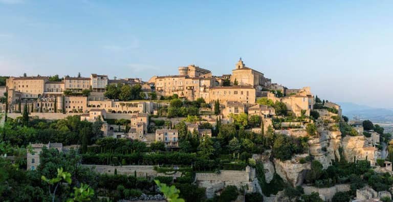 Hilltop village of Gordes with stone buildings and terraced gardens, surrounded by greenery and airelles, under a clear blue sky.