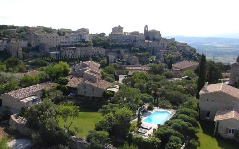 A hillside village like Beaume or Gordes with stone buildings, green gardens, and a swimming pool, overlooking a scenic valley under a bright sky.