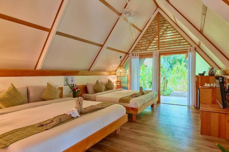 A tropical-style room at Shawandha Lodge in Puerto Viejo, Costa Rica, with two double beds, wooden floors, decorative towels, a small desk area, and glass doors leading to a lush garden outside.