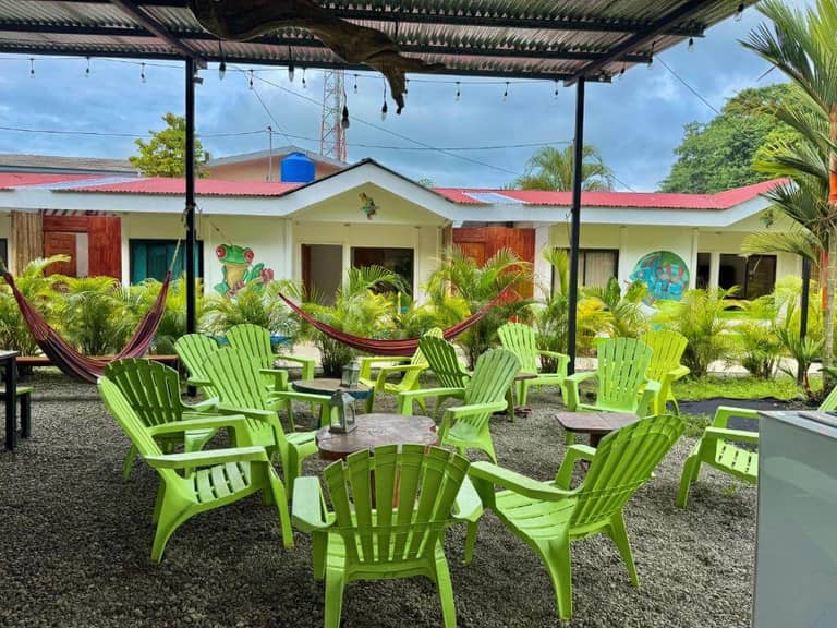 Outdoor patio at Kalunai Hostel in Puerto Viejo, featuring green plastic chairs, round tables, and hammocks under a covered area, surrounded by lush plants and vibrant murals on the building in the background.