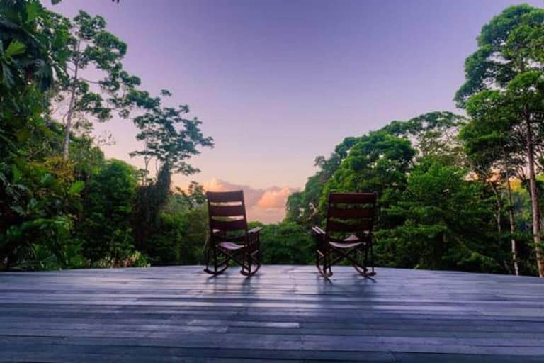 Two wooden rocking chairs sit on a deck overlooking a lush green forest with a clear sky at sunrise or sunset.